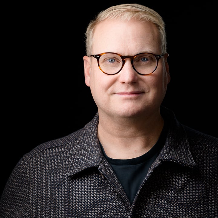 Tim Lockie headshot wearing a collared grey pattern shirt with a black background